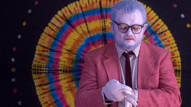 old mime in big glasses with blue hair and beard in bright pink jacket and tie in white gloves. clown rotates his hands and makes magic passes. Hand training before performance. Happy clown