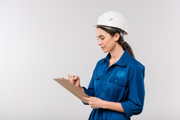 Young successful female engineer in blue workwear and safety helmet making notes