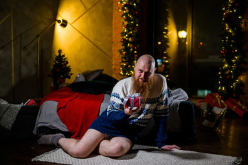 Obraz na płótnie Canvas A man with a long red beard sits on the floor without pants in a winter sweater. man is holding a wrapped present on the background of New Year decorations and lights. Christmas tree. Parody, humor.