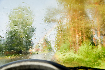 View from car cabin on wet asphalt road, trees and rainbow during rain and drops on front window