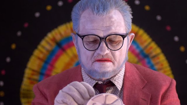 elderly mime man in large glasses with blue hair and beard in bright pink jacket and tie in white gloves. clown does makeup on face. White face, blue hair, red lips. Happy clown