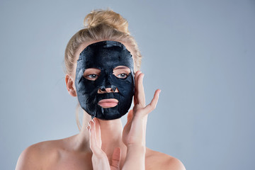 girl with naked shoulders posing at grey background with facial mask black, skin care concept, beauty photo, skin treatment, hydrating skin mask, smiling.