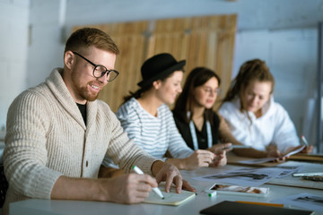 Creative fashion designer drawing sketch in copybook on background of colleagues