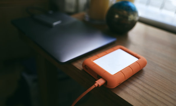 An External Hard Drive Backup Device Connected To A Laptop On A Wooden Table With A Blurred Background.