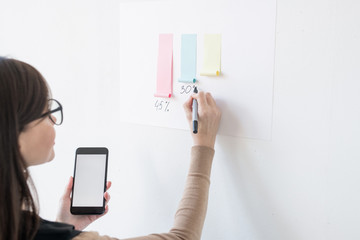 Young female broker writing down percentage under paper chart on whiteboard