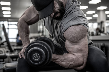 strong young bearded man in cap lifting heavy weight dumbbell on muscle biceps sitting with great effort and pain during hard workout training in sport gym