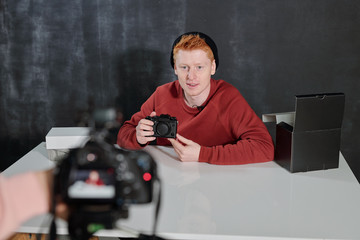 Young man in casualwear sitting by desk and talking about new photocamera
