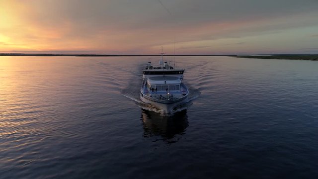 Flight around cinematic modern cruise ship boat vessel floats on calm water. People tourists on deck. Lena river lake Siberia Russia. Epic orange sunset clouds natural landscape. Open space horizon