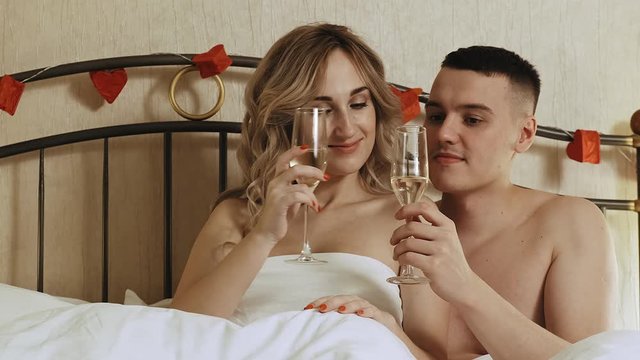 A young couple sits in bed, under a blanket. They're choking on champagne glasses and drink. Romantic date in a hotel room