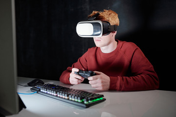 Young man with gamepad in vr headset sitting in front of computer screen