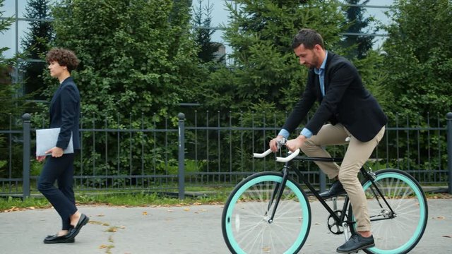 Healthy adult man is riding bicycle to work in city street full of businesspeope walking to office building, green trees are visible in background. Youth and health concept.