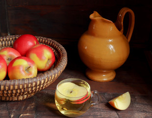 A cup of apple juice or apple cider, a jug and fresh colourful red and yellow apples apples in a wicker plate on the dark old wooden background