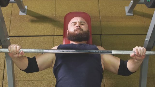 Handsome brutal powerlifter enjoying bodybuilding training in fitness club, lying on red bench with heavy barbell in hands