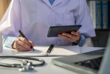 Female medicine doctor, physician or practitioner in lab room writing on blank notebook and work on laptop computer with medical stethoscope on the desk at hospital. Medic tech concept.