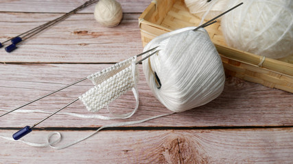 hands knitting white color from bamboo threads, needles and knitted thing in the process of knitting next to a wicker box with balls of yarn on a wooden table
