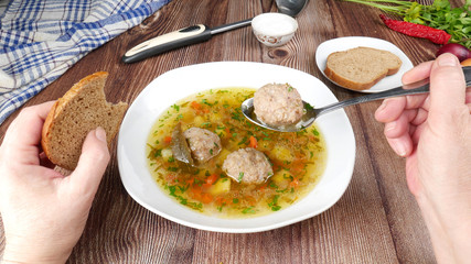 Soup with vegetables and meatballs in a white plate on a wooden table. Female hands holding a spoon with soup and a slice of dark bread