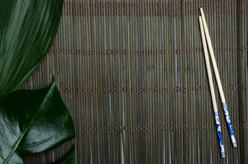 Top view of dark green bamboo mat, fresh exotic leaves and wooden chopsticks.Empty space for design