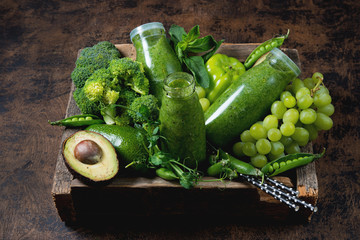 Three bottles of the healthiest green smoothie from vegetables and fruits are in a wooden box and around the green vegetables and fruits. Horizontal photo.