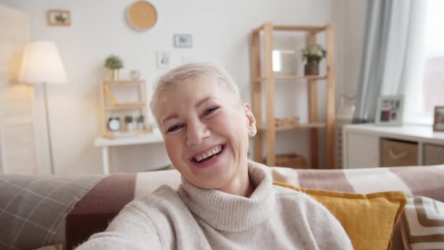 Chest-up shot of 50-something Caucasian lady with cropped grey hair sitting on couch at home, holding invisible smartphone in outstretched hand, waving and chatting enthusiastically on video call
