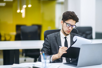 Young office worker in glasses reads documents in the modern office. Young business manager working with new startup project. Stylish man in a suit reviews and analyze documents, plans.