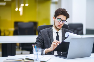 Young office worker in glasses reads documents in the modern office. Young business manager working with new startup project. Stylish man in a suit reviews and analyze documents, plans.