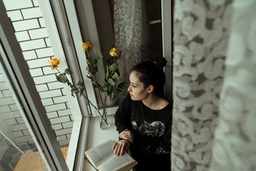 A girl in black clothes sits by the open window and reads a book. Yellow roses are standing nearby.
