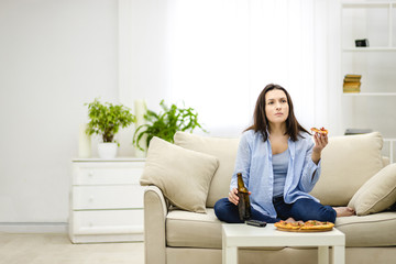 Brunette woman chews, holding a bottle of beer and a slice of pizza hands. Isolated woman on blurred light background. Copy space.