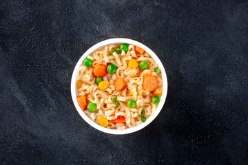 Ramen cup, instant noodles in a plastic cup, with green peas and carrot, shot from the top with copy space on a black background