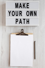 'Make your own path' words on a modern board, clipboard with blank sheet of paper on a white wooden surface, top view. Overhead, from above, flat lay. Copy space.