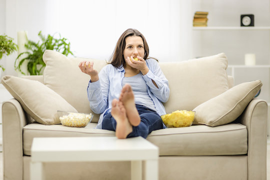 Portrait of pretty, charming, cheerful woman with straight black hair, who is eating pop corn and potato chips, watching comic serial, comedy, sitting with crossed legs in living room.