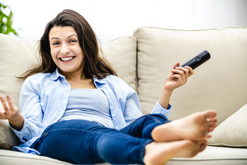 Happy young woman is watching tv, sitting on a sofa. She is smiling widely. Copy space.