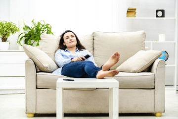 Obraz premium Bored and relaxed woman is watching TV, laying on a sofa. Copy space.