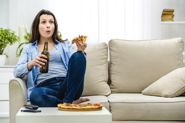 Attractive and interested woman is shocked, watching television. Amazed woman is sitting on the lisht sofa and eating snacks.