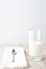 Glass of milk with metal spoon on white napkin on wooden table. High key