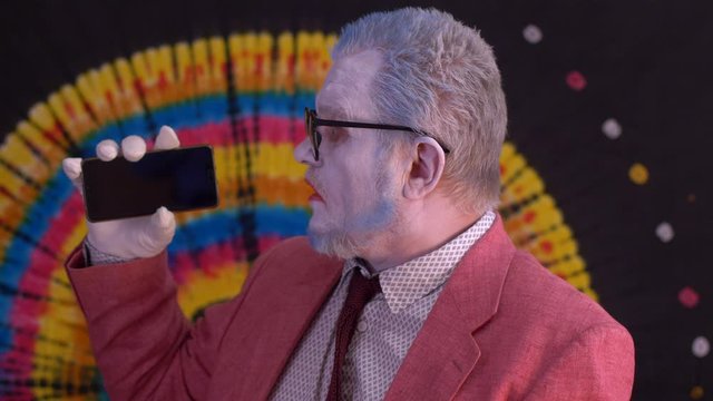 elderly mime man in big glasses with blue hair and beard in bright pink jacket and tie looks at smartphone screen and shows it to audience. Laughter, tears, positive and negative emotions. Happy clown