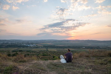 Obraz premium Young man sits with his samoyed dog among the field and enjoying amazing landscape with sunset