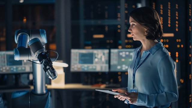 Female Engineer With Tablet Computer Analyse How A Futuristic Robotic Arm Works And Moves A Metal Object. High Tech Research Laboratory With Modern Equipment.