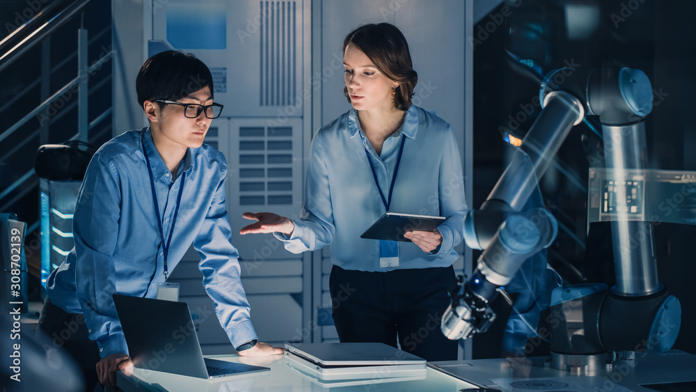 Fototapete man and woman engineers with laptop and a tablet analyse and discuss how a futuristic robotic arm wo