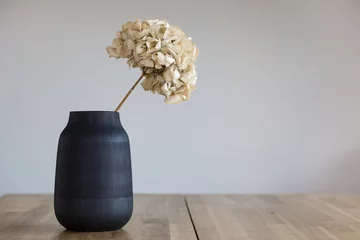 Tuinposter Een zwarte keramische vaas met een gedroogde hortensia bloem staat op een houten ondergrond. © papa