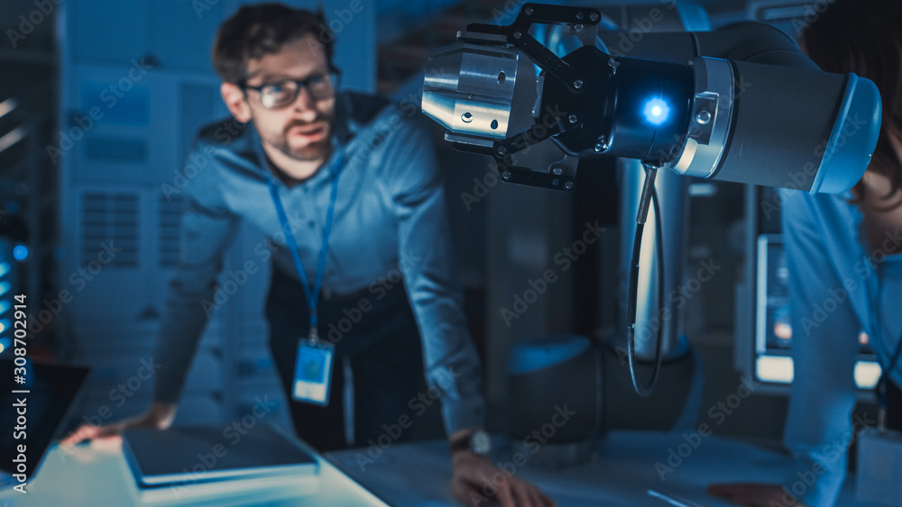 Poster Close Up of a Futuristic Robotic Arm Picking Up a Metal Object and Moving It. Team of Engineers Following This Advanced Process. They are in a High Tech Research Laboratory with Modern Equipment.