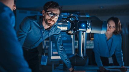 Futuristic Metal Robotic Arm Picking Up a Metal Object and Moving It. Senior Engineer Talks Through This Advanced Process. They are in a High Tech Research Laboratory with Modern Equipment.