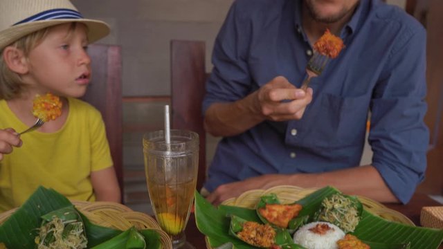 Young man and his little son eat traditional Balenese food set Nasi Campur in a traditional cafe. Travel to Bali concept