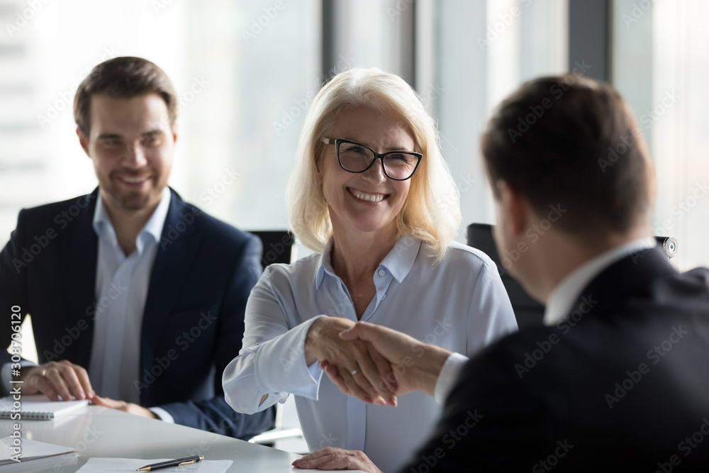 Poster Smiling mature businesswoman shaking hand of business partner at meeting