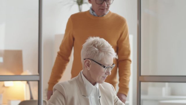 Medium shot of mature Caucasian man and woman with grey hair, in glasses working together in small office, looking at computer screen and discussing business matters