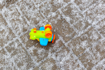 Fototapeta na wymiar A children's, full-color, plastic airplane lies on the gray stone pavement covered with snowflakes of the first December snow.