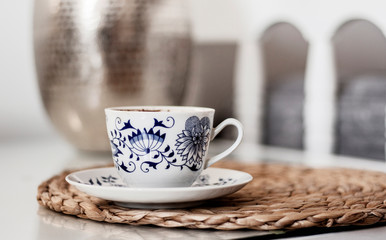 Cup of coffee on wicker stand on a glass white table, metal vase on the background, cozy home interior concept.