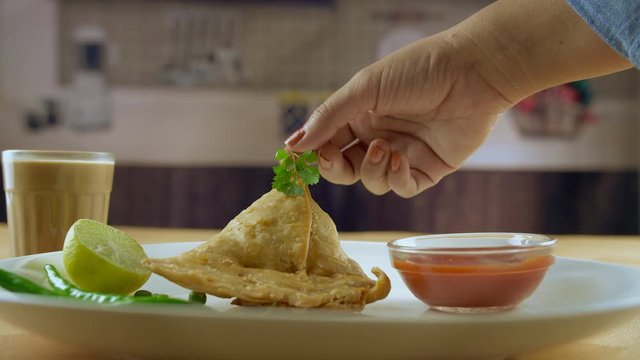 Hands of a woman decorating samosa plate with a coriander leaf at home in India. Beautifully plated samosa with a bowl of tomato sauce  sliced lemon  green chilies  coriander leaf and masala chai(tea)