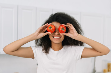 Happy emotion African American woman holding red tomatoes near eyes, standing  in the kitchen, having fun. Healthy lifestyle concept, vegetarian, diet. Attractive emotional girl cooking fresh salad 