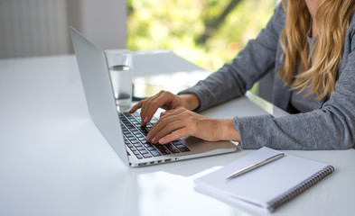 A blonde hair businesswoman working in her workstation. Businesswoman working at the desk at workplace. Woman working in home office. Woman sitting at desk and working with laptop and glass of water.