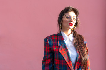 Waist up portrait of the young attractive brunette femalee in glasses, with red lips, dressed in plaid jacket and white shirt, isolated over pink background